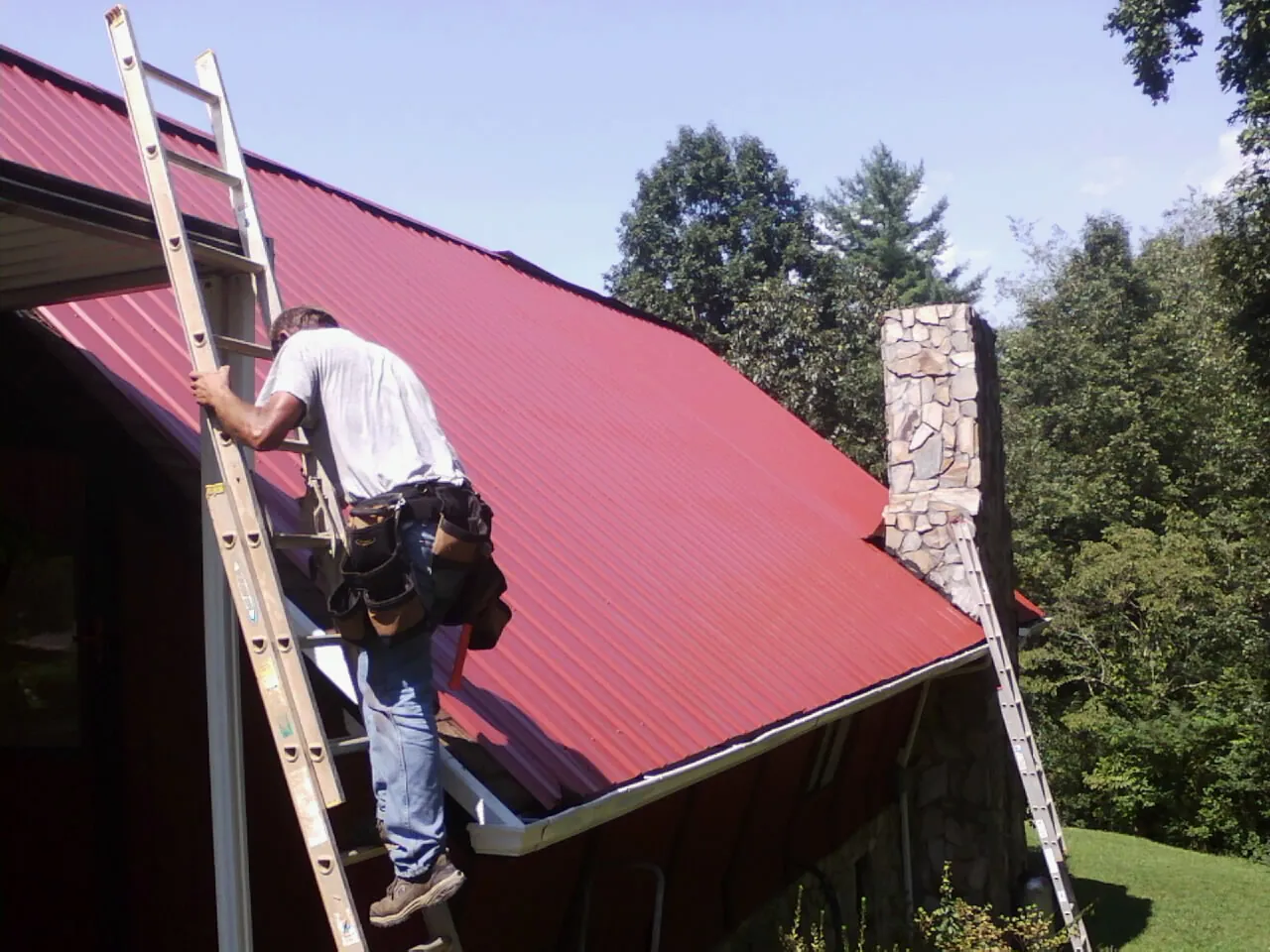 Roofer on a ladder beside a red metal roof and stone chimney — Soffit & Fascia Repair by Roofing Brothers in Fitzgerald